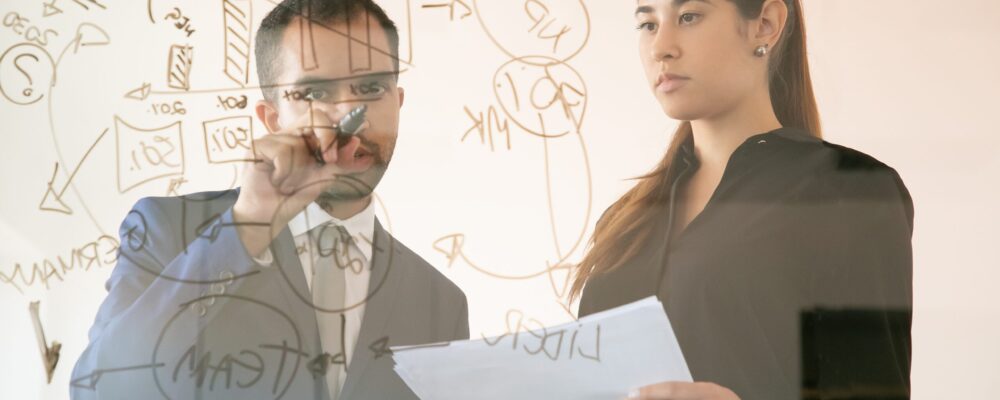 Content African American manager writing chart on glass board. Professional young pretty female colleague holding document and looking at graph in conference room. Teamwork and marketing concept